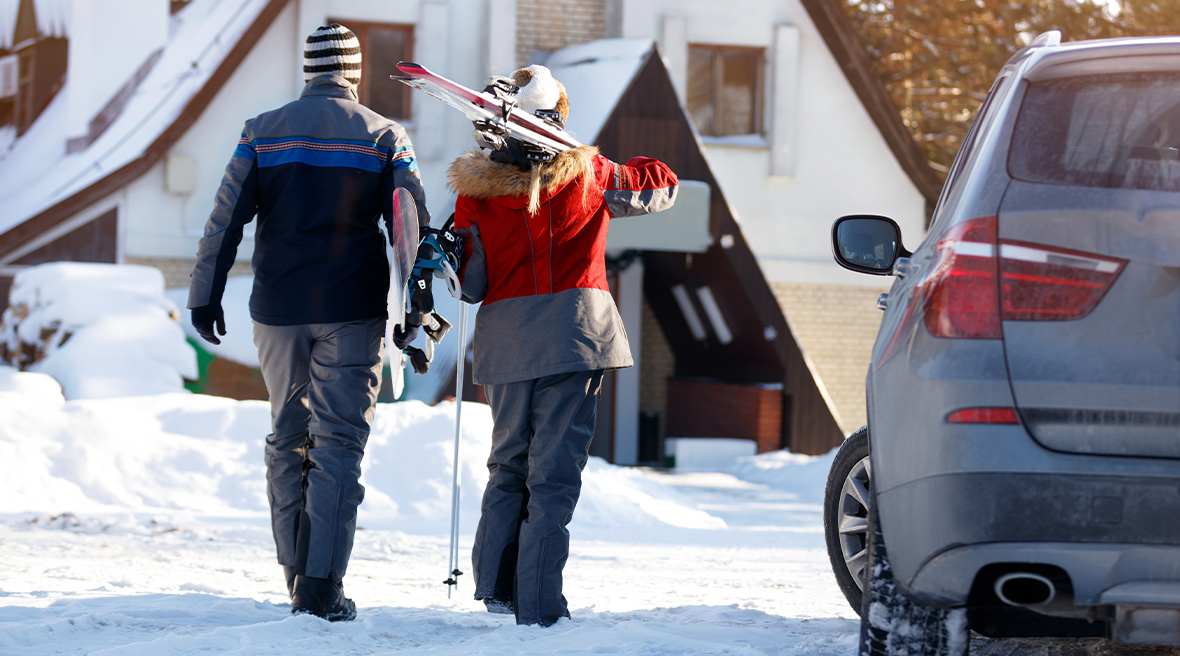 couple carrying skis and ski poles from parked car to wooden chalet in the snow